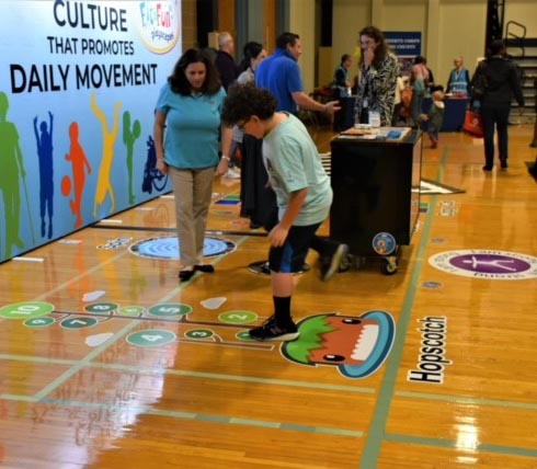 On April 29th, 2023 Dutchess County Department of Behavioral & Community Health hosted their 2nd Annual YOUR Health Fair at Dutchess Community College’s Falcon Hall. Fit and Fun Playscapes were present with a large area where they demonstrated many of their popular activity stickers to keep individuals active and engaged while. Here a young person engages in Hopscotch, while owner, Pam looks on.