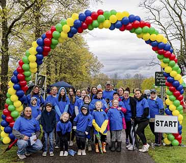 A team of about 20 individuals of all ages and abilities, dressed in blue t-shirts over hoodies and jackets, are under the rainbow colored balloon arch. 2023 Autism Directory Services Walk for Autism at James Baird Park on Saturday April 29, 2023