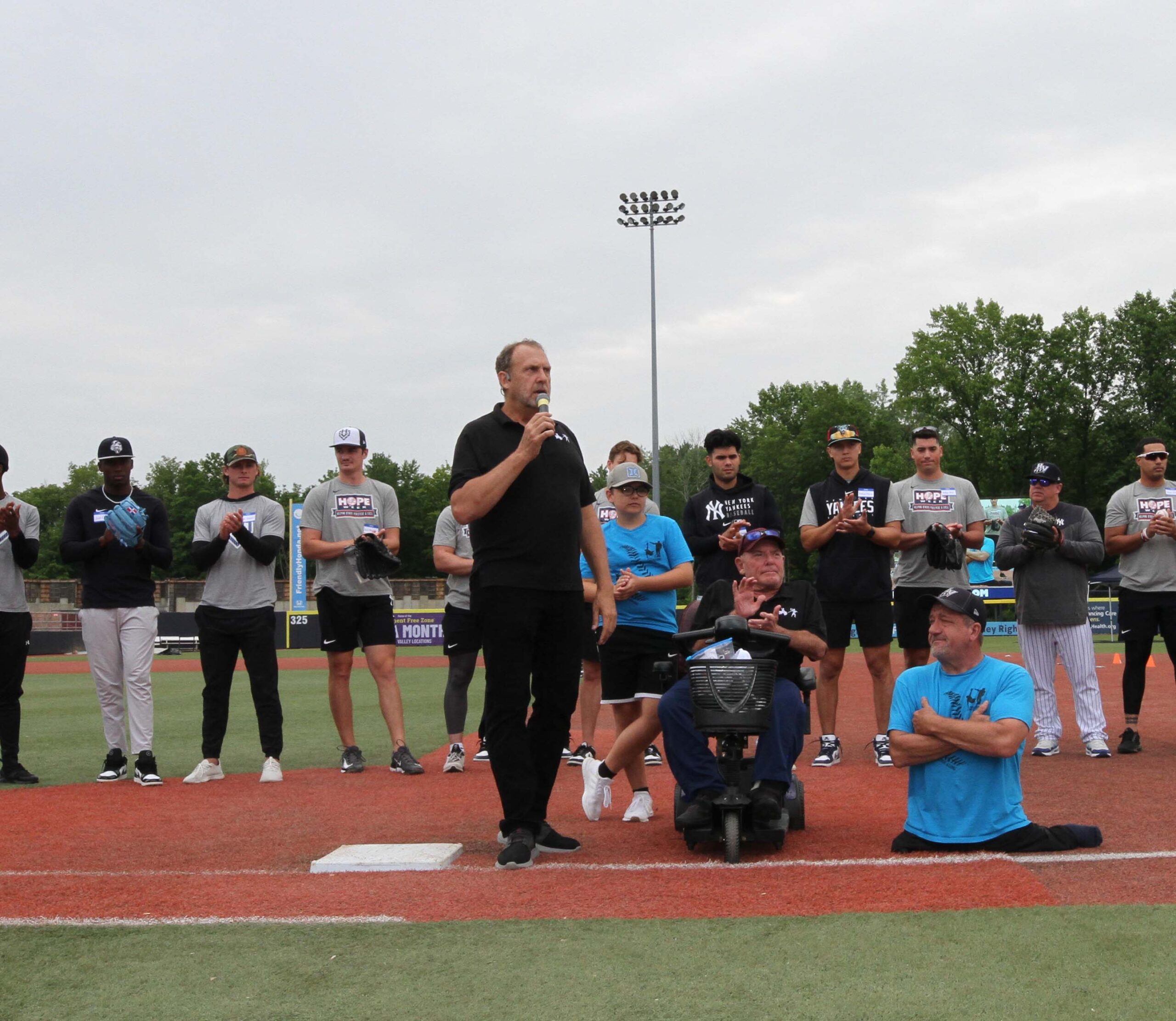 Doug Cornfield of The Dave Clark Foundation and Pulling Each Other Along, addresses the crowd of the D-3 Day with former MLB Pitcher Dave Clark (seated on an electric scooter) to his right, and Dave Stevens, to the far right, sitting/standing on the ground. Behind the gentlemen are members of the Hudson Valley Renegades. Disability, Dream and Do (also known as D3 Day) at Heritage Financial Field, June 17, 2023.