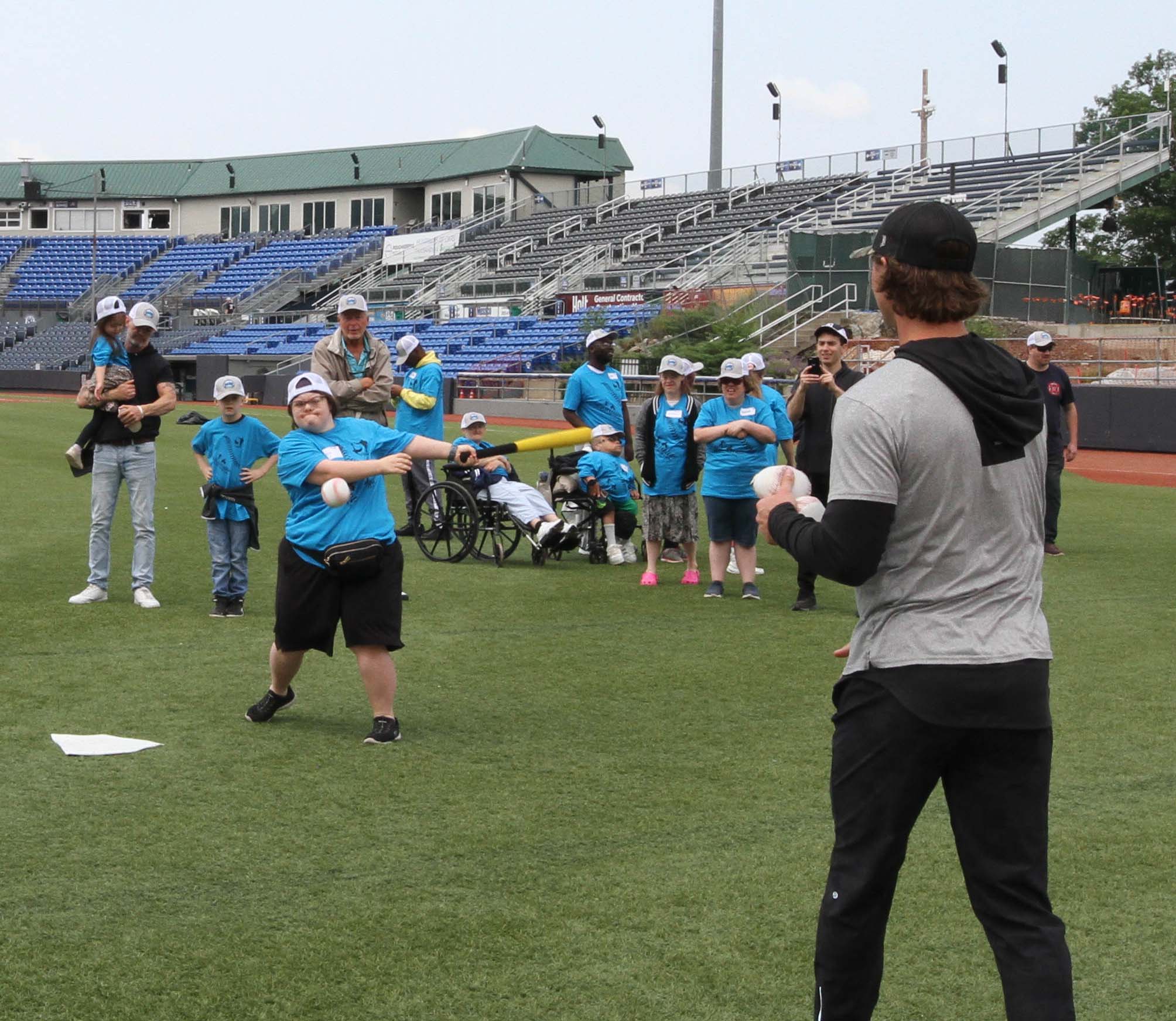 A participant, with a white baseball hat turned backwards, hits a baseball thrown by a Hudson Valley Renegades Player (their back is to us). Behind the hitter a group of participants awaits their turn to hit a baseball. Disability, Dream and Do (also known as D3 Day) at Heritage Financial Field, June 17, 2023