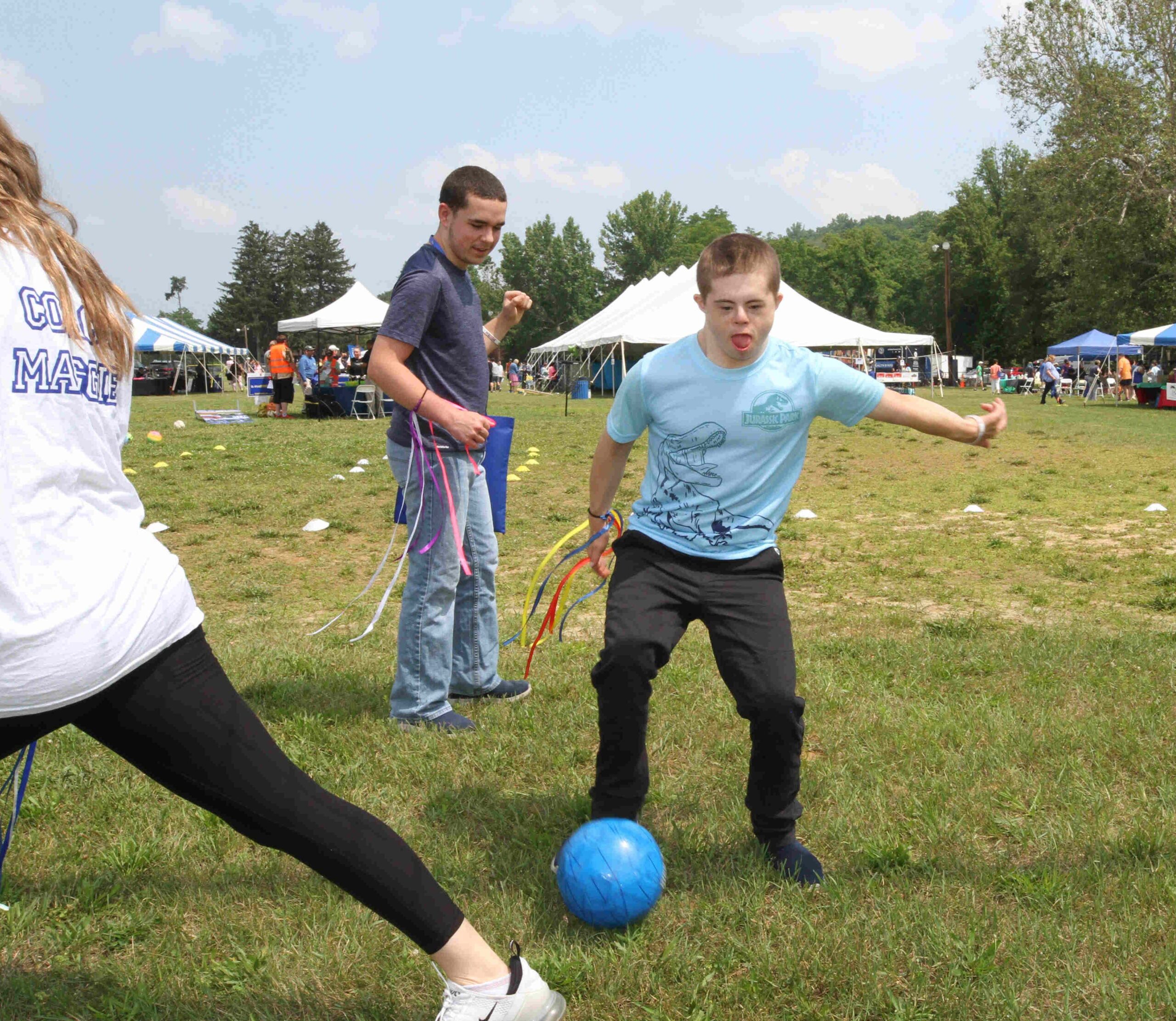 2 young people play soccer with Coach Maggie (who’s back is to us in the picture). Both of them are wearing colorful ribbons around their wrists, another activity that was found at the 3rd Annual ThinkDIFFERENTLY Fitness & Field Day on Friday June 29, 2023 at Bowdoin Park.