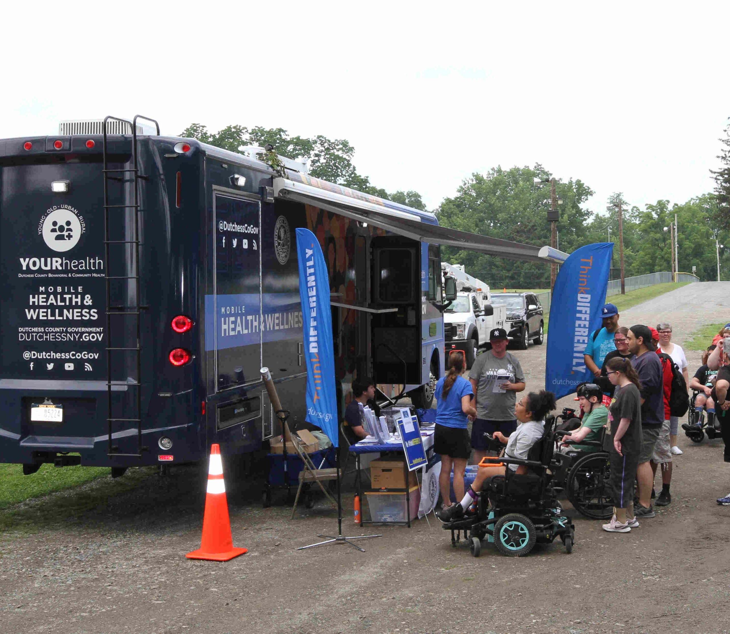 The Dutchess County Mobile Health & Wellness RV was on hand to provide information, resources, check in, and RV tours at the 3rd Annual ThinkDIFFERENTLY Fitness & Field Day on Friday June 29, 2023 at Bowdoin Park. In this picture the large 40’ long Navy Blue shows off the back and passenger side details, which include white writing noting “YOUR Health (Young, Old, Urban, Rural), Dutchess County Government Dutchessny.gov and social media handle of @dutchesscogov. Along the passenger side of the RV about 15 people stand around the tables of resources and check in for the ThinkDIFFERENTLY Fitness and Field Day.